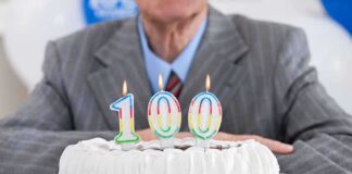 A man in a suit sitting in front of a birthday cake with 100 candles