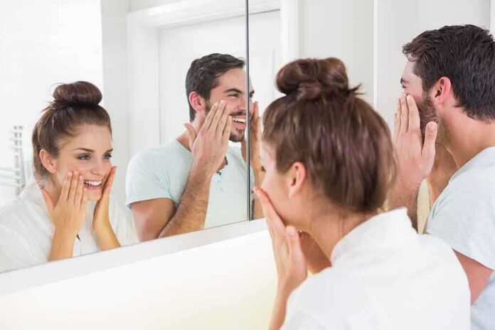 A couple smiling at their reflections in a bathroom mirror while applying skincare products