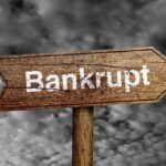 Wooden arrow sign pointing towards the word Bankrupt against a cloudy background