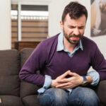 Terrifying Vomiting Wave Slams The US Man sitting on couch, holding stomach in pain.