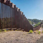 Holiday Immigration Rant Sparks Media Fury Tall metal border wall with rural landscape.