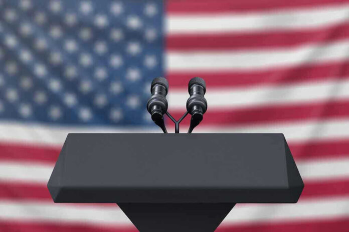 Podium with microphones, American flag background.