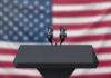 Podium with microphones, American flag background.