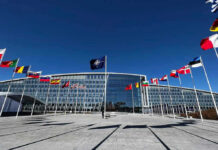 U.S. Pullback JOLTS NATO Plans Flags outside NATO headquarters building under clear blue sky.