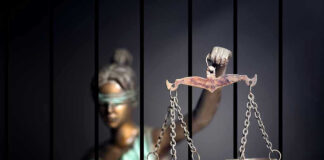 Blindfolded Lady Justice statue holding scales behind bars.