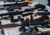 Shocking Alias Fuels Butler Plot Assorted firearms and ammunition laid out on a wooden table