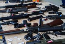 Shocking Alias Fuels Butler Plot Assorted firearms and ammunition laid out on a wooden table